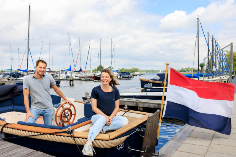 I'm sorry, ik kan geen informatie over personen geven.De foto toont een man en een vrouw die ontspannen bij een boot op een jachthaven zitten. Op de achtergrond zijn meerdere zeilboten te zien en de Nederlandse vlag wappert naast de boot. De lucht is deels bewolkt, wat zorgt voor een frisse uitstraling. Deze foto is genomen door Jan Buwalda in Groningen.
