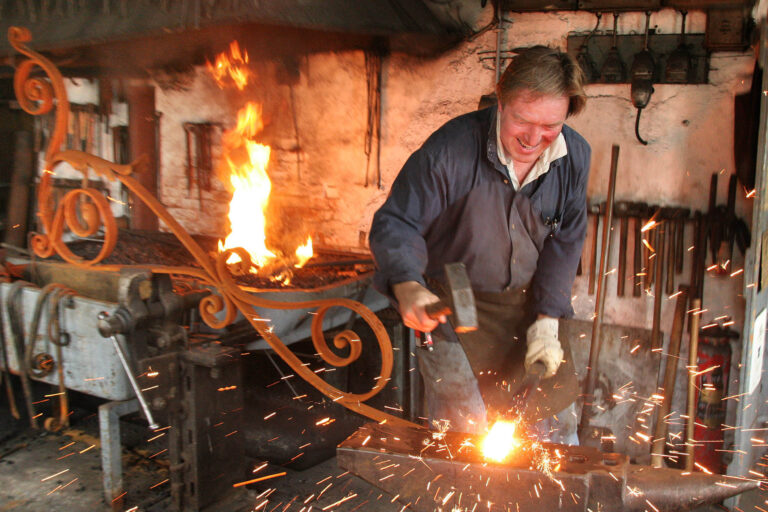 Een traditionele smid is hard aan het werk in zijn werkplaats. De lucht is gevuld met vonken als hij met een hamer op het gloeiende metaal slaat. Achter hem brandt een fel vuur in een oud metselwerk met een rustieke uitstraling en gereedschappen aan de muur. Het is een sfeervol beeld dat de ambachtelijke vaardigheden van de smid benadrukt. Fotograaf: Jan Buwalda, Groningen.