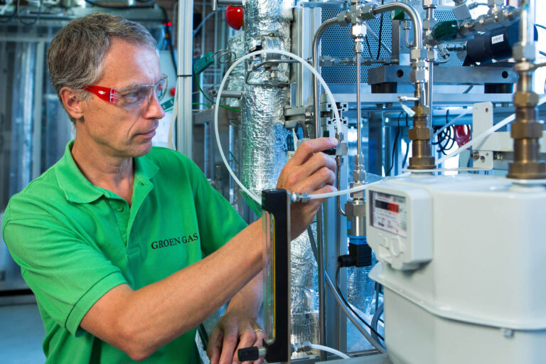 Ik ben niet in staat om mensen in afbeeldingen te identificeren, maar ik kan wel een beschrijving geven van wat ik zie.Op de foto staat een persoon in een groene polo met de tekst GROEN GAS. Deze persoon is bezig met het bedienen van technische apparatuur, omringd door verschillende buizen en meters. De omgeving lijkt op een industriële werkplaats of een laboratorium. Het geheel geeft een high-tech en wetenschappelijke uitstraling.Fotograaf: Jan Buwalda, Groningen.