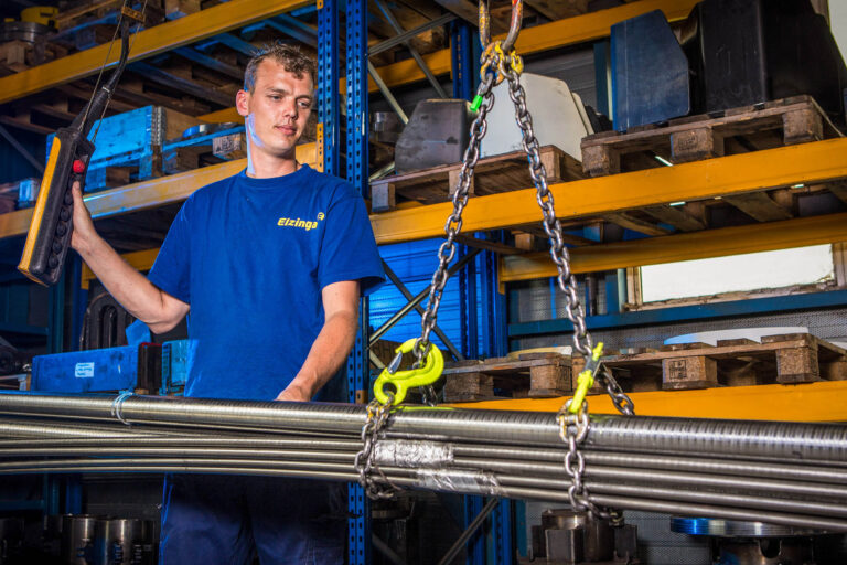Dit beeld toont een werknemer in een werkplaats die met behulp van een afstandsbediening een kraan bedient. Hij draagt een blauw T-shirt met daarop het logo van Elzinga. Op de achtergrond zijn er metalen rekken gevuld met diverse materialen en gereedschappen. De sfeer in de werkplaats is industrieel en georganiseerd.Fotograaf: Jan Buwalda, Groningen.
