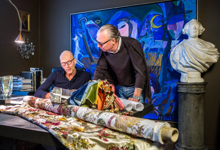 Ik kan helaas de namen van mensen op de foto niet bevestigen.Op de foto zijn twee mannen te zien in een creatieve werkruimte. Ze bespreken stoffen en ontwerpen, omringd door kleurrijk kaastrelen en een imposante buste. Een artistieke sfeer heerst in de kamer. Fotografie door Jan Buwalda in Groningen.