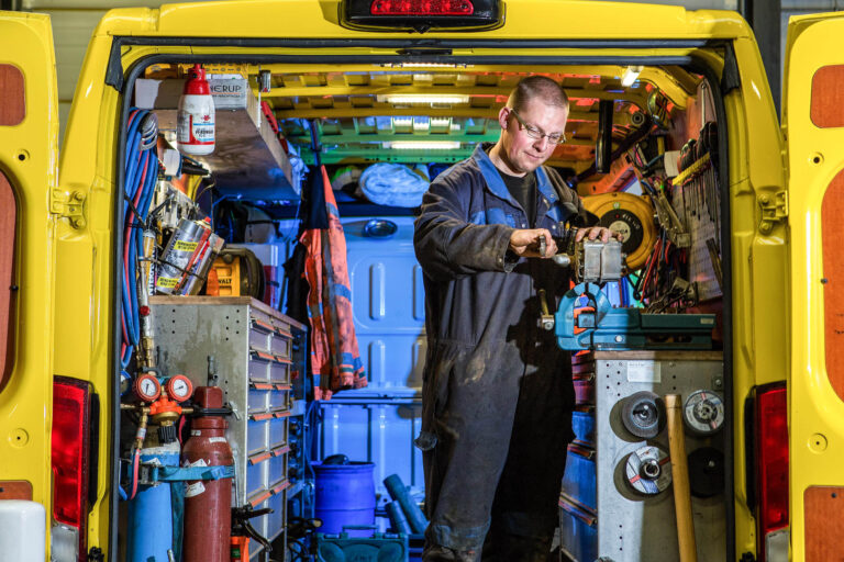 Dit is een foto van het interieur van een werkbus, waarin een man druk bezig is met een reparatiesessie. De bus is uitgerust met diverse gereedschappen en apparatuur die zorgvuldig geordend zijn voor optimaal gebruik. De man draagt werkkleding en werkschoenen en lijkt geconcentreerd bezig met een montage in een bankschroef. De goed georganiseerde opstelling van de bus wijst op een efficiënte werkplek voor mobiele reparaties. Fotograaf: Jan Buwalda, Groningen.