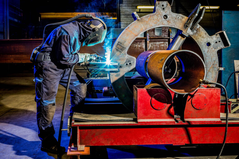 Dit beeld toont een lasser in een industriële omgeving bezig met het lassen van grote metalen onderdelen. De lasser draagt beschermende kleding, inclusief een lashelm, en wordt omgeven door een sterke blauwe gloed van het lasproces. De werkplek is gevuld met zware apparatuur en metalen constructies, wat de technische en industriële sfeer benadrukt. Foto door Jan Buwalda, Groningen.