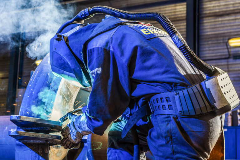 Dit is een indrukwekkende foto van een lasser aan het werk, genomen door Jan Buwalda in Groningen. De lasser draagt een beschermend pak en een helm met luchtfiltersysteem, wat essentieel is voor de veiligheid tijdens het lassen. De vonken spatten in het rond terwijl de lasser geconcentreerd aan het werk is. De belichting van de foto benadrukt de details van de kleding en de apparatuur, en vangt de dynamiek van het lasproces op een krachtige manier.