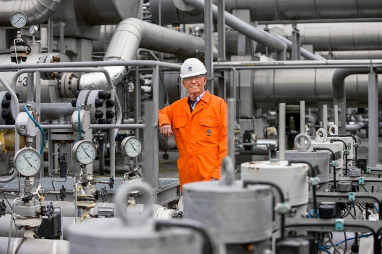 Ik kan de persoon op de foto niet identificeren, maar de afbeelding toont een persoon in een oranje overall en witte veiligheidshelm, poserend te midden van complexe industriële pijpleidingen en meters. Dit lijkt een gas- of oliefaciliteit te zijn, met veel metalen buizen en technische apparatuur. De foto straalt een gevoel van technische expertise en industriële kracht uit. Fotograaf: Jan Buwalda, Groningen.