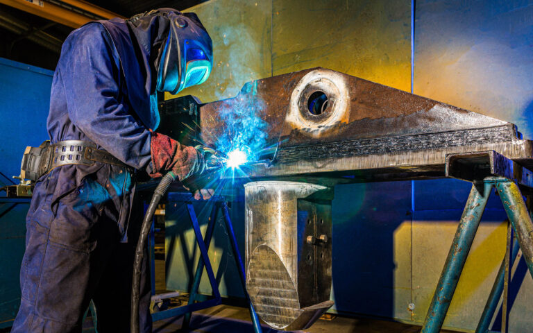 Een lasser is geconcentreerd aan het werk in een werkplaats, met een vlamboog die helder oplicht terwijl hij een groot metalen onderdeel bewerkt. De vonken springen in de rondte en creëren een dynamisch beeld tegen de industriële achtergrond. De beschermende kleding en laskap beschermen de lasser tijdens het precisiewerk. De foto vangt de intensiteit en vakbekwaamheid van het lasproces. Fotograaf: Jan Buwalda, Groningen.