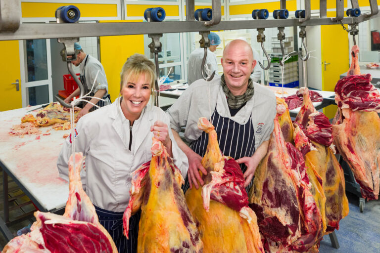 In een helder verlichte slagerij staan twee medewerkers met een glimlach te midden van opgehangen stukken vlees. Ze dragen witte jassen en strepen schorten. Op de achtergrond zijn andere werknemers bezig met het bewerken van vlees op werktafels. De ruimte heeft gele accenten aan de muren. Foto door Jan Buwalda in Groningen.