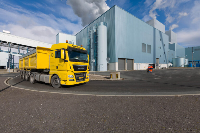 Dit beeld toont een grote, felgele vrachtwagen met de merknaam MAN, die voor een industriële fabriek rijdt. De fabriek is modern, met een lichtblauwe gevel en verschillende silo's en industriële buizen zichtbaar aan de zijkant. Boven de fabriek stijgt witte rook op tegen de blauwe lucht met enkele wolken. Op de achtergrond zijn twee personen in feloranje veiligheidshesjes te zien, wat op een industriële werkomgeving duidt. Dit alles is vastgelegd door fotograaf Jan Buwalda in Groningen.