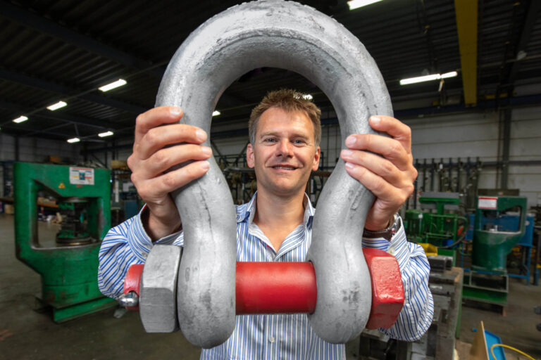 Dit is een foto van een man in een werkplaats, die een grote metalen sluiting vasthoudt. Hij glimlacht naar de camera te midden van industriële machines en apparatuur, die een achtergrond vormen van zware industrie en techniek. De foto legt de nadruk op de robuuste bouw van de sluiting en de industriële omgeving. Fotograaf: Jan Buwalda, Groningen.
