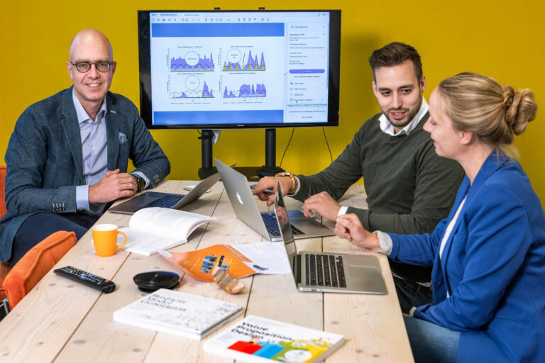 Ik kan de personen op de foto niet identificeren, maar ik kan wel een beschrijving geven van de situatie. De afbeelding toont drie mensen aan een vergadertafel, druk in overleg. Allen hebben laptops voor zich en er ligt leesmateriaal op de tafel. Op de achtergrond is er een beeldscherm met grafieken zichtbaar. De gele muur voegt een frisse tint toe aan de ruimte. Deze foto is gemaakt door Jan Buwalda in Groningen.