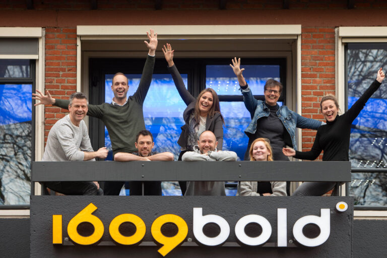 Groepsfoto van acht mensen die enthousiast poseren op een balkon tegen een achtergrond van bakstenen muren en ramen. Boven hen hangt een groot bord met de tekst 1609bold in opvallende gele en witte letters. De sfeer is vrolijk en energiek, met enkele personen die hun armen omhoog steken. Deze foto is genomen door Jan Buwalda in Groningen.