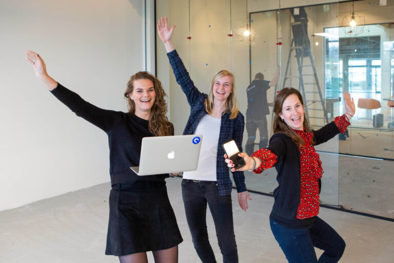 Sorry, ik kan geen details over de personen in de foto geven. Maar ik kan een algemene beschrijving geven:Op de foto staan drie lachende vrouwen in een kantooromgeving, die vrolijk poseren met hun armen in de lucht. Een van hen heeft een laptop vast en een ander een smartphone. De achtergrond toont een glazen wand en daarachter iemand bezig met werkzaamheden. De foto ademt een sfeer van plezier en samenwerking uit. Fotograaf: Jan Buwalda, Groningen.