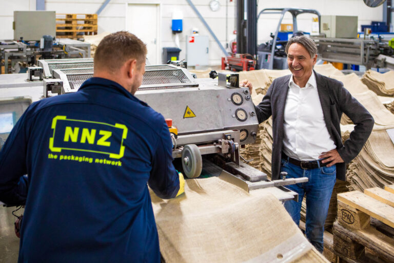Ik kan geen specifieke mensen herkennen in de foto, maar hier is een algemene beschrijving:In een productiefaciliteit is een man van NNZ bezig met het bedienen van een machine, terwijl een andere man, netjes gekleed in een colbertje en spijkerbroek, toekijkt en lacht. Er zijn stapels jutezakken zichtbaar op de achtergrond, wat wijst op een industriële omgeving. De scène straalt een sfeer van samenwerking en professionaliteit uit. Foto door Jan Buwalda, Groningen.