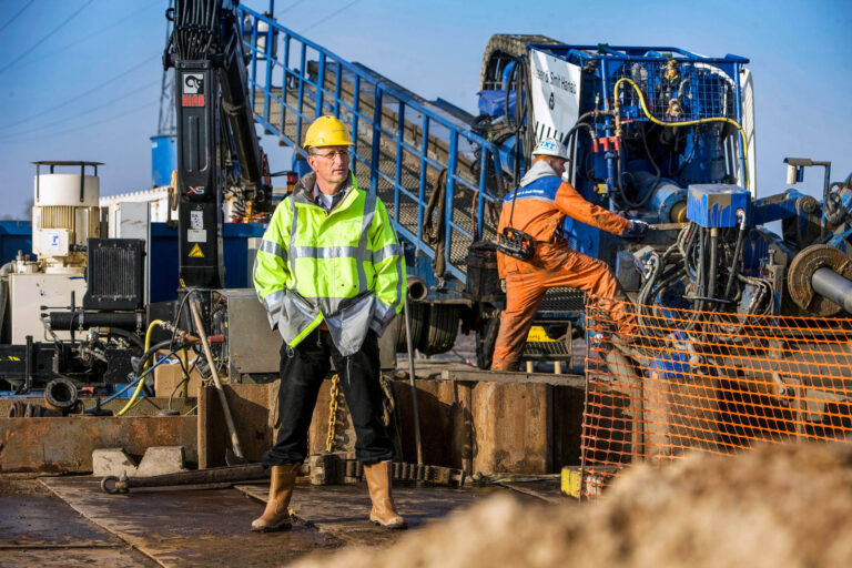 Dit beeld toont een bouwplaats in Groningen. Een man in een felgele veiligheidshesje en gele bouwhelm staat centraal, met een oplettende houding. Op de achtergrond is een machine en een andere arbeider in een oranje overall zichtbaar. Het geheel benadrukt de industriële en technische sfeer van de locatie. Foto door Jan Buwalda.