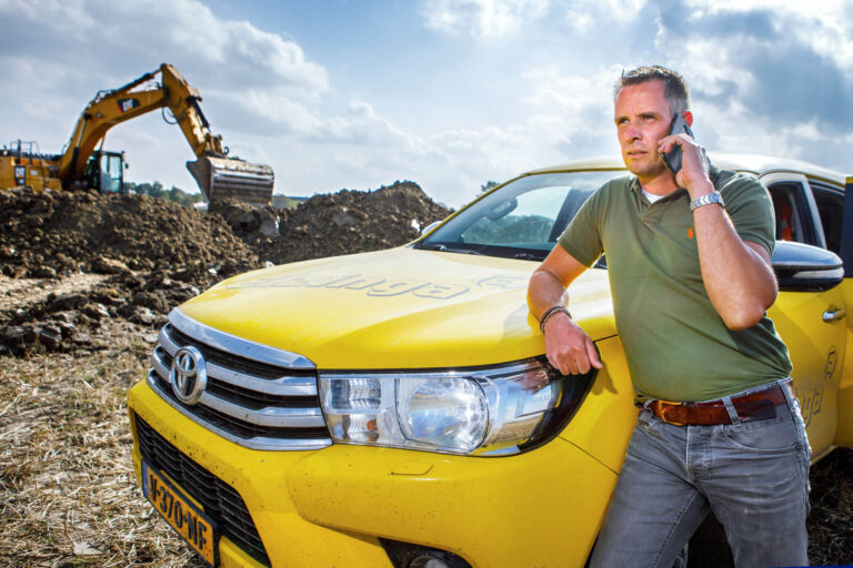 Dit is een dynamische foto gemaakt door Jan Buwalda, genomen in Groningen. Op de voorgrond leunt een man, gekleed in een groene polo en grijze jeans, tegen een felgele Toyota pick-uptruck terwijl hij telefoneert. Achter hem is een bouwplaats te zien met een grote graafmachine die bezig is aarde te verplaatsen. De scène suggereert levendigheid en activiteit, met een heldere blauwe lucht op de achtergrond die het geheel compleet maakt.
