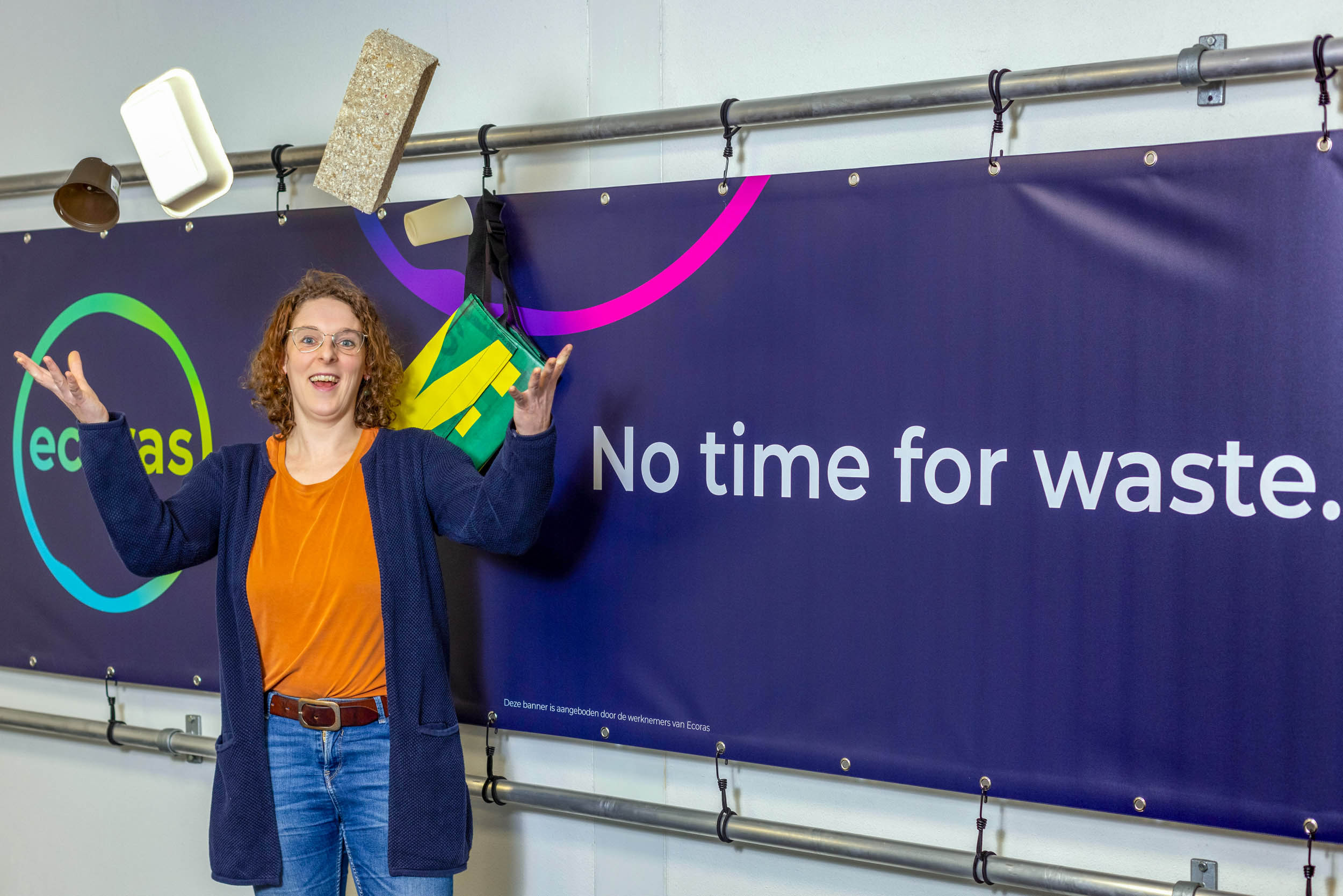 In deze foto, genomen door Jan Buwalda, zien we een persoon in een moderne ruimte voor een kleurrijke banner met de tekst No time for waste. De persoon lijkt enthousiast en betrokken bij het onderwerp. Rondom haar zweven verschillende alledaagse voorwerpen in de lucht, wat een dynamisch en speels effect geeft aan de compositie. De achtergrond en de attributen benadrukken mogelijk een thema van duurzaamheid en efficiënt gebruik van middelen. De foto is gemaakt in Groningen.