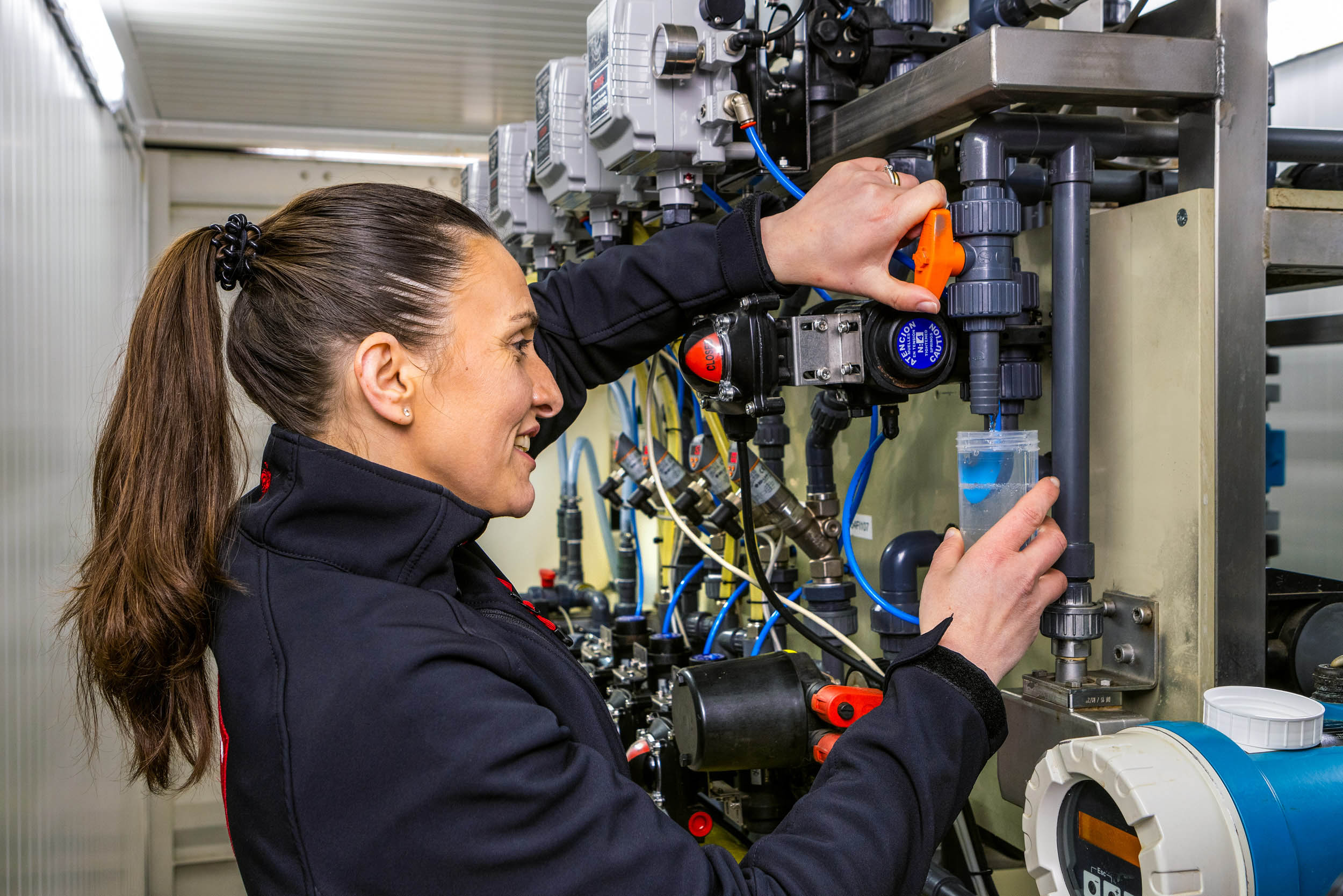 Ik kan de persoon in de foto niet identificeren, maar ik kan wel een algemene beschrijving geven.Deze foto toont een vrouw die bezig is met het bedienen van industriële apparatuur. Ze draagt een donkere jas en haar haar is in een staart gebonden. Ze is geconcentreerd bezig met een onderdeel van de installatie, waarbij ze een oranje hendel bedient en een doorzichtige container houdt waarin een blauwe vloeistof wordt opgevangen. De omgeving lijkt op een technische ruimte met diverse leidingen en apparatuur.Fotograaf: Jan Buwalda, Groningen.