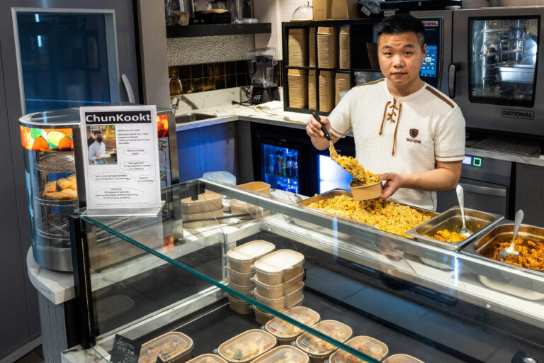 Een kok staat achter de toonbank van een keuken, bezig met het opscheppen van rijst in een kartonnen bakje. Voor de toonbank staan verschillende verpakkingen met etenswaren, en links is een bord te zien met informatie over ChunKookt. De keuken is modern ingericht met diverse kookapparatuur. Fotograaf: Jan Buwalda, Groningen.