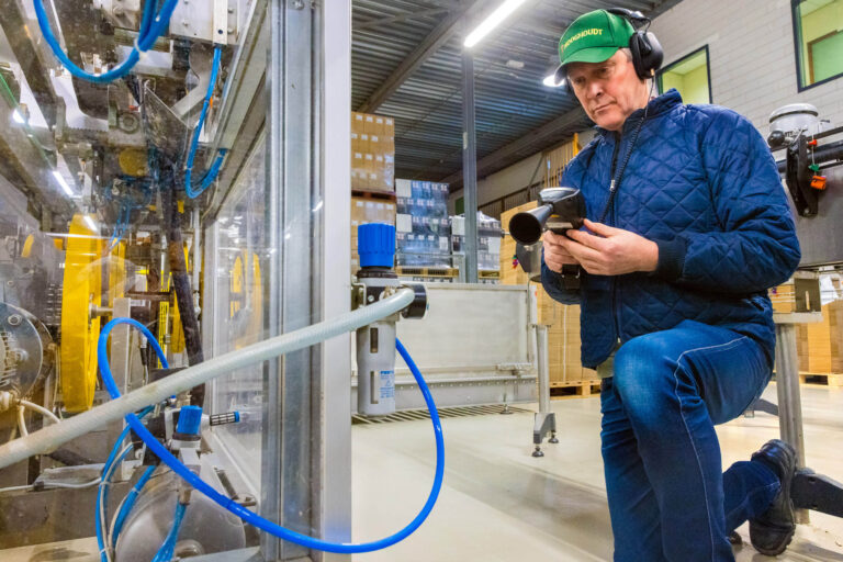 Een medewerker knielt bij een industriële machine in een fabriek. Hij draagt een groene pet en gehoorbescherming en bedient een handscanner. Op de achtergrond zijn dozen en apparaten zichtbaar, wat wijst op een georganiseerde productieomgeving. De foto is genomen door Jan Buwalda in Groningen.