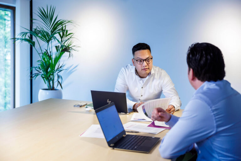 Deze foto toont een zakelijke setting waarbij twee mensen aan een vergadertafel zitten. Ze zijn in gesprek en hebben documenten en laptops voor zich op tafel liggen, wat wijst op een professionele bespreking. Op de achtergrond staat een grote groene plant, wat een natuurlijke toets aan de ruimte geeft. Het geheel presenteert een moderne en lichte kantooromgeving. Fotograaf: Jan Buwalda, Groningen.