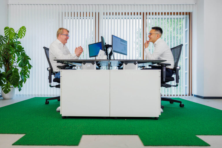 Een gedetailleerde foto van een moderne kantoorruimte waarin twee mannen tegenover elkaar aan een groot bureau zitten. Beiden dragen nette, witte overhemden en lijken in een geanimeerd gesprek verwikkeld. Op de achtergrond zijn verticale witte lamellen te zien die natuurlijk licht binnenlaten, en er staat een grote groene plant links. De vloer is bedekt met een groen tapijt dat de ruimte een frisse uitstraling geeft. De foto is gemaakt door Jan Buwalda in Groningen.
