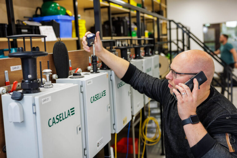 Een man in een werkplaats is bezig met een Casella-apparaat aan de muur. Hij lijkt gegevens te nemen met een kleine sensor terwijl hij met iemand telefoneert. Op de achtergrond staan verschillende gereedschappen en apparatuur. De ruimte heeft een georganiseerde en professionele sfeer. Fotograaf: Jan Buwalda, Groningen.