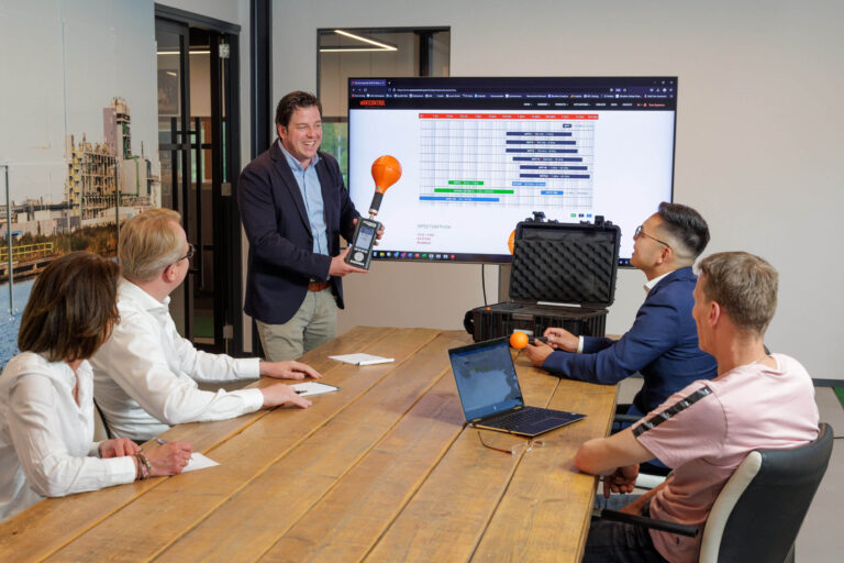 Deze afbeelding toont een zakelijke bijeenkomst waarbij vijf mensen aan een grote houten tafel zitten. Eén persoon staat en presenteert een apparaat met een oranje bol aan de bovenkant. Op de achtergrond is een grote monitor zichtbaar, waarop een planningsschema staat afgebeeld. De ruimte lijkt modern en professioneel, met een industrieel ogend gebouw zichtbaar op de wanddecoratie. De deelnemers aan het gesprek lijken geïnteresseerd en aandachtig. Foto door Jan Buwalda, Groningen.