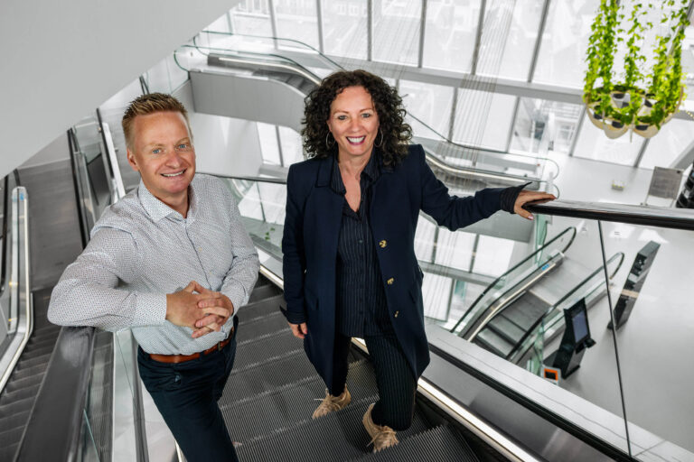 Dit is een foto van twee mensen op een roltrap in een modern gebouw met veel glas en lichtinval. Op de achtergrond zijn grote ramen zichtbaar die uitzicht geven op de stad. De man draagt een licht overhemd en de vrouw een donkere jas. Ze kijken allebei vriendelijk naar de camera. De groene planten die van het plafond hangen geven een fris accent aan de ruimte. Fotografie door Jan Buwalda, Groningen.