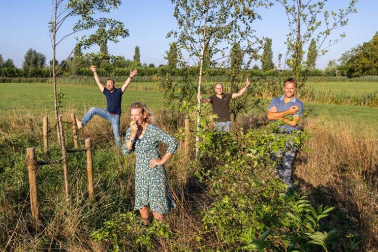 Dit is een levendige foto genomen door Jan Buwalda in Groningen. Op de foto staan vier personen midden in een groene omgeving met jonge bomen. De vrouw op de voorgrond draagt een jurk met een patroon en neemt een hap van een stuk fruit. Een man op de achtergrond springt enthousiast in de lucht, terwijl een andere man met een glimlach tegen een boom leunt met een appel in zijn arm. De vierde persoon staat ontspannen bij een boom, zijn handen omhoog. De foto straalt een gevoel van vreugde en verbondenheid met de natuur uit.