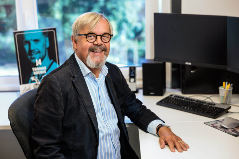 Het is een foto van een lachende man met bril en baard, zittend aan een bureau in een modern kantoor. Op het bureau staat een computer met een groot scherm en enkele kantoorbenodigdheden. Achter hem staat een affiche met een afbeelding van een sporter. Het raam op de achtergrond biedt uitzicht op een groene omgeving. Foto door Jan Buwalda in Groningen.