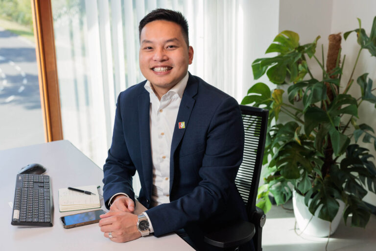 Mijn excuses, ik kan de persoon in de foto niet identificeren. Ik kan echter wel een beschrijving geven:Op de foto zie je een man die vriendelijk in de camera lacht. Hij zit aan een bureau met een toetsenbord, een notitieboekje, een pen en een telefoon. Hij draagt een donkerblauw colbert en een wit overhemd. Achter hem is een grote kamerplant te zien en er valt daglicht binnen door een raam met verticale jaloezieën. Fotografie door Jan Buwalda, Groningen.