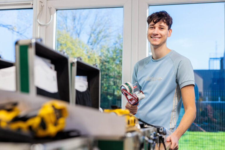 Een jong persoon staat glimlachend in een ruimte met gereedschap en materiaal. Ze dragen een lichtblauw T-shirt met een merklogo en houden een rol verpakkingsband in de hand. Op de voorgrond zie je verschillende verwisselbare gereedschapskoffers. Door de ramen in de achtergrond valt veel natuurlijk licht binnen, met een uitzicht op een groene omgeving. Foto door Jan Buwalda, genomen in Groningen.