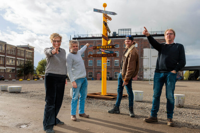 Het beeld toont vier personen die enthousiast poseren voor een geel richtingsbord op een industrieterrein met historische bakstenen gebouwen op de achtergrond. De lucht is deels bewolkt, wat zorgt voor een helder en levendig contrast. De sfeer is ontspannen en vrolijk. Foto door Jan Buwalda in Groningen.