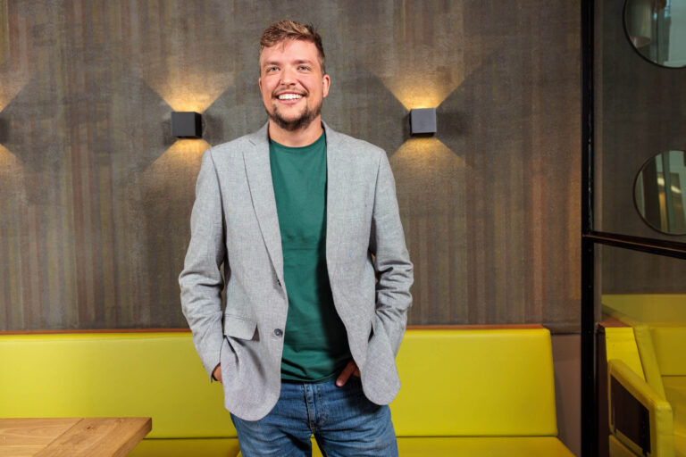 Het is een portretfoto van een lachende man met een baard, gekleed in een lichtgrijs colbert en een groene trui, gecombineerd met een spijkerbroek. Hij staat ontspannen met zijn handen in zijn zakken voor een achtergrond met een modern patroon en sfeerverlichting. De foto geeft een professionele en toegankelijke uitstraling. Fotograaf: Jan Buwalda, Groningen.