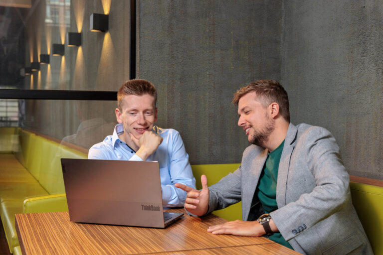 Twee mannen zitten aan een houten tafel in een moderne, stijlvolle ruimte. Ze kijken geïnteresseerd naar het scherm van een laptop voor hen. De man links draagt een lichtblauw overhemd, terwijl de man rechts een grijs jasje over een groen T-shirt draagt. De verlichting langs de muur draagt bij aan een warme en professionele sfeer. Foto door Jan Buwalda in Groningen.