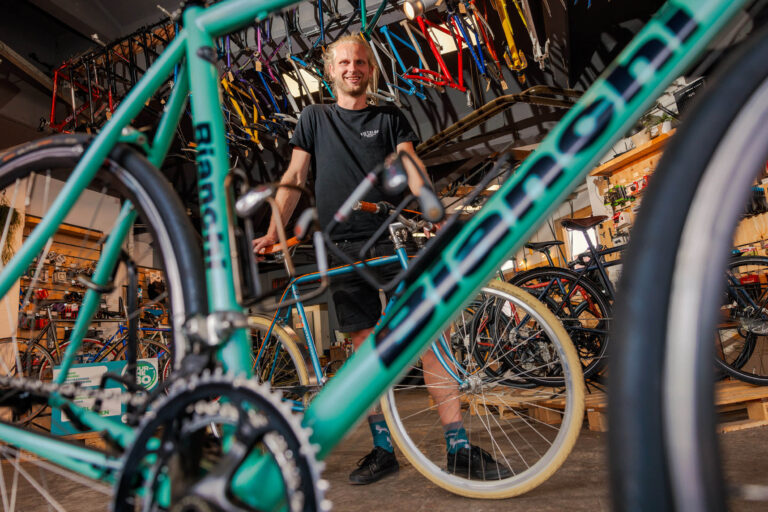 Het beeld toont een persoon in een fietsenwinkel, omringd door een diverse collectie fietsen. Op de voorgrond is een groene Bianchi fiets prominent in beeld. De winkel heeft een industriële uitstraling met fietsonderdelen aan de muur. De persoon straalt betrokkenheid en passie voor fietsen uit. Fotograaf: Jan Buwalda, Groningen.