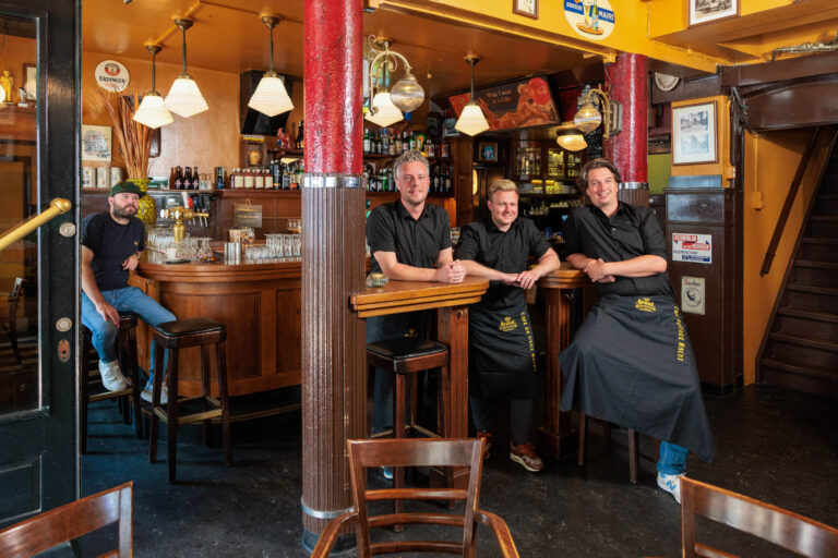 Dit sfeervolle en authentieke café in Groningen toont vier mannen ontspannen poseren in een gezellige en warme omgeving. Drie mannen, gekleed in zwarte bedieningsuniformen met schorten, leunen tegen de houten bar of staan dichtbij, terwijl een andere man links van hen aan de bar zit. De inrichting straalt een klassieke bruine kroeguitstraling uit met houten details, uitnodigende verlichting en decoratieve elementen. De foto is genomen door de fotograaf Jan Buwalda.