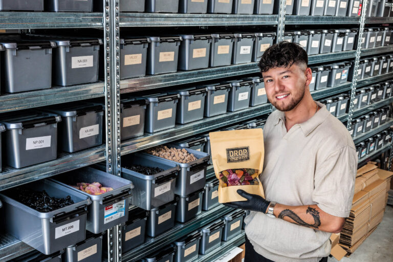 Een jongeman staat glimlachend voor een stellingkast gevuld met grijze bakken die netjes zijn geëtiketteerd. Hij draagt een lichtgekleurde polo en zwarte handschoenen, en houdt een papieren zak met de tekst DROP vegan erin. De ruimte straalt een georganiseerde en professionele sfeer uit, en de bakken bevatten verschillende soorten drop en snoepgoed. Fotograaf: Jan Buwalda, Groningen.