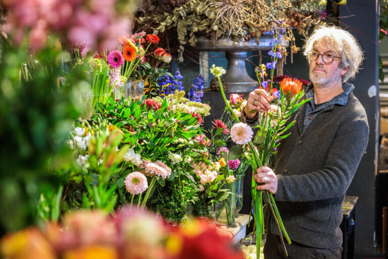 Een man staat in een bloemenwinkel omringd door een kleurrijke selectie van bloemen. Hij houdt een boeket in zijn hand en lijkt zorgvuldig bloemen te schikken. Op de achtergrond zijn diverse soorten bloemen zichtbaar, waaronder gerbera's en andere seizoensbloemen. De scène straalt een gevoel van creativiteit en toewijding uit. Deze foto is gemaakt door Jan Buwalda in Groningen.