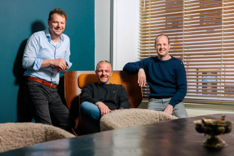 Dit is een foto van drie mannen in een moderne en comfortabele kantooromgeving. De man links staat met zijn hand op een stoel, gekleed in een lichte blouse en donkere broek. In het midden zit een man ontspannen op een stijlvolle houten stoel met een zwart shirt en blauwe jeans. Rechts, leunt een man op dezelfde stoel, gekleed in een donkerblauwe trui en lichte broek. De ruimte is verlicht door natuurlijk licht dat door de houten jaloezieën naar binnen valt.Fotograaf: Jan Buwalda, Groningen.