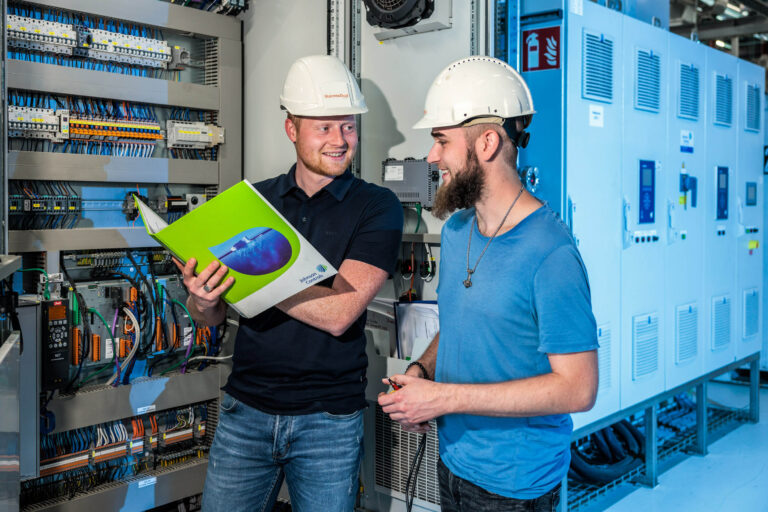 Twee mannen in werkkleding en met veiligheidshelmen staan voor een grote schakelkast vol met bedrading en componenten. De ene man houdt een kleurrijke map vast en lacht terwijl hij met de andere man praat. Deze foto is genomen door Jan Buwalda in Groningen.
