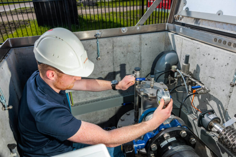 Dit beeld toont een man die aan het werk is in een technisch station. Hij draagt een veiligheidshelm en een donkerblauwe polo, terwijl hij aan een installatie werkt met verschillende meetinstrumenten en leidingen. De omgeving lijkt op een industriële ruimte met metalen en betonnen oppervlakken. Het licht valt helder op de scène, wat de nadruk legt op de details van zijn werkzaamheden. Foto door Jan Buwalda, Groningen.