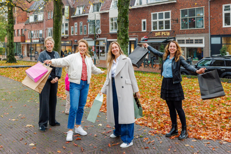 Een levendige foto genomen in een pittoreske straat in Groningen tijdens de herfst. Vier vrolijke vrouwen staan midden op een met bladeren bezaaid trottoir, elk met kleurrijke winkelzakken in hun handen. Ze stralen enthousiasme uit, te midden van een charmante achtergrond met bakstenen gebouwen en een café genaamd De Pub. De warme herfstkleuren van de vallende bladeren voegen een gezellige sfeer toe aan de scène. Fotografie door Jan Buwalda.