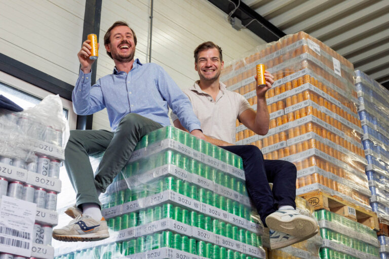 Dit is een foto genomen in een magazijn waar twee lachende mannen opgestapelde pallets met blikjes drinken zitten. Ze houden blikjes vast en kijken blij. Achter hen zijn er meerdere stapels pallets met verschillende kleuren blikjes te zien. De sfeer lijkt ontspannen en vrolijk. Fotografie door Jan Buwalda in Groningen.
