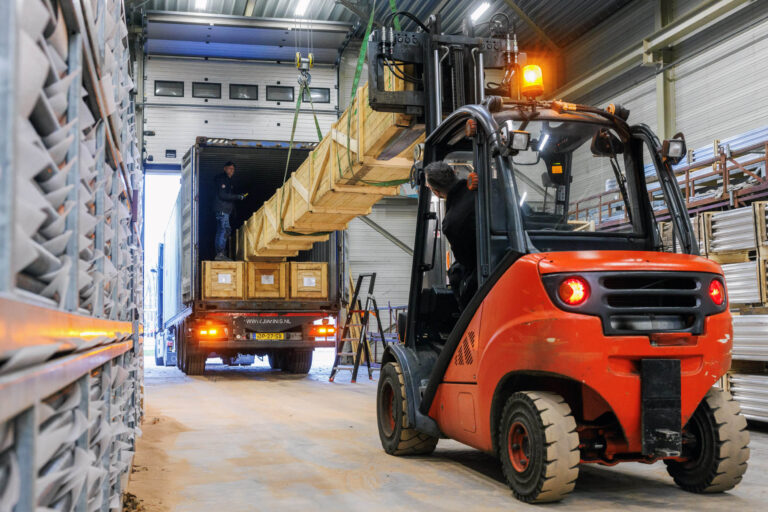 Dit beeld toont een logistieke scène in een magazijn in Groningen. Op de voorgrond is een heftruckchauffeur bezig met het verplaatsen van een lange houten kist, terwijl een vrachtwagen op de achtergrond geladen wordt. Het magazijn is gevuld met rekken vol materiaal en er speelt een dynamische workflow. De verlichting en de open deur zorgen voor voldoende lichtinval. Fotograaf: Jan Buwalda.