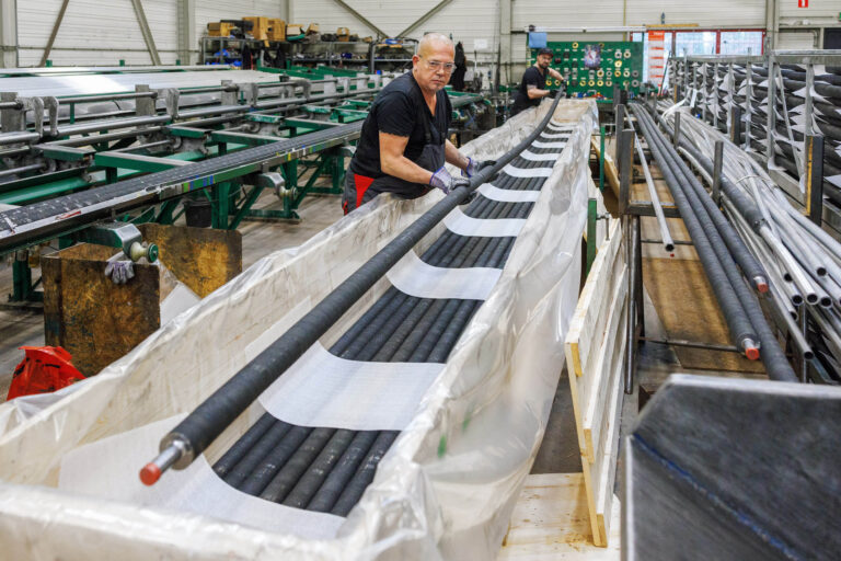 In deze industriële scène is een werknemer bezig in een fabriekshal waar lange buizen worden verwerkt. Hij draagt veiligheidsbrillen en handschoenen terwijl hij nauwkeurig met de materialen werkt. Op de achtergrond is een collega bezig met soortgelijke werkzaamheden. De omgeving is gevuld met metalen onderdelen en productieapparatuur. Fotograaf: Jan Buwalda, Groningen.