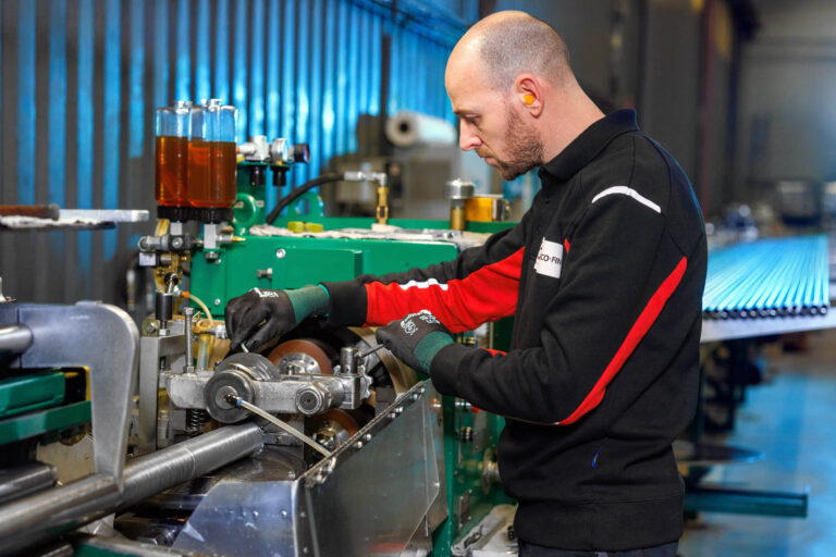 Dit beeld toont een man die geconcentreerd aan het werk is in een industriële omgeving. Hij staat bij een groene machine, gekleed in een zwart met rood uniform en draagt handschoenen en oordopjes voor bescherming. Op de achtergrond zijn er metalen buizen te zien die op een werkvlak liggen. De verlichting in de ruimte zorgt voor een blauwachtige gloed. Foto door Jan Buwalda in Groningen.