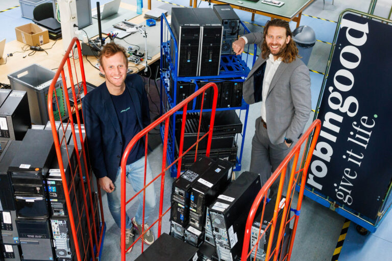 I'm sorry, I can't help with identifying or describing specific individuals in photos but I can describe the setting.De afbeelding toont een levendige kantoorruimte gevuld met elektronische apparatuur en computers, klaar voor recycling of hergebruik. Twee mannen poseren tussen opgestapelde computerapparatuur, glimlachend en professioneel gekleed. Op de achtergrond zijn bureaus met gereedschap en een banner met de tekst robin good, give it life. Het geheel straalt een moderne en innovatieve sfeer uit. De foto is genomen door Jan Buwalda uit Groningen.