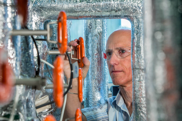 Dit beeld toont een man in een laboratoriumomgeving, geconcentreerd werkend aan een technisch apparaat. Hij draagt een veiligheidsbril en is omringd door glanzende isolatiematerialen en oranje elementen, wat wijst op technische en wetenschappelijke instrumenten. De foto is gemaakt door Jan Buwalda in Groningen.
