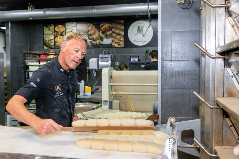 Dit beeld toont een bakker aan het werk in een moderne bakkerij. De bakker legt deegvormen op een bakplaat, klaar om gebakken te worden. Op de achtergrond zijn verschillende broden en bakkersoorten te zien op een bord. De omgeving lijkt schoon en professioneel, met apparaten en gereedschappen die klaarstaan voor gebruik. Het logo van de bakkerij is zichtbaar op de muur. Fotografie door Jan Buwalda, Groningen.